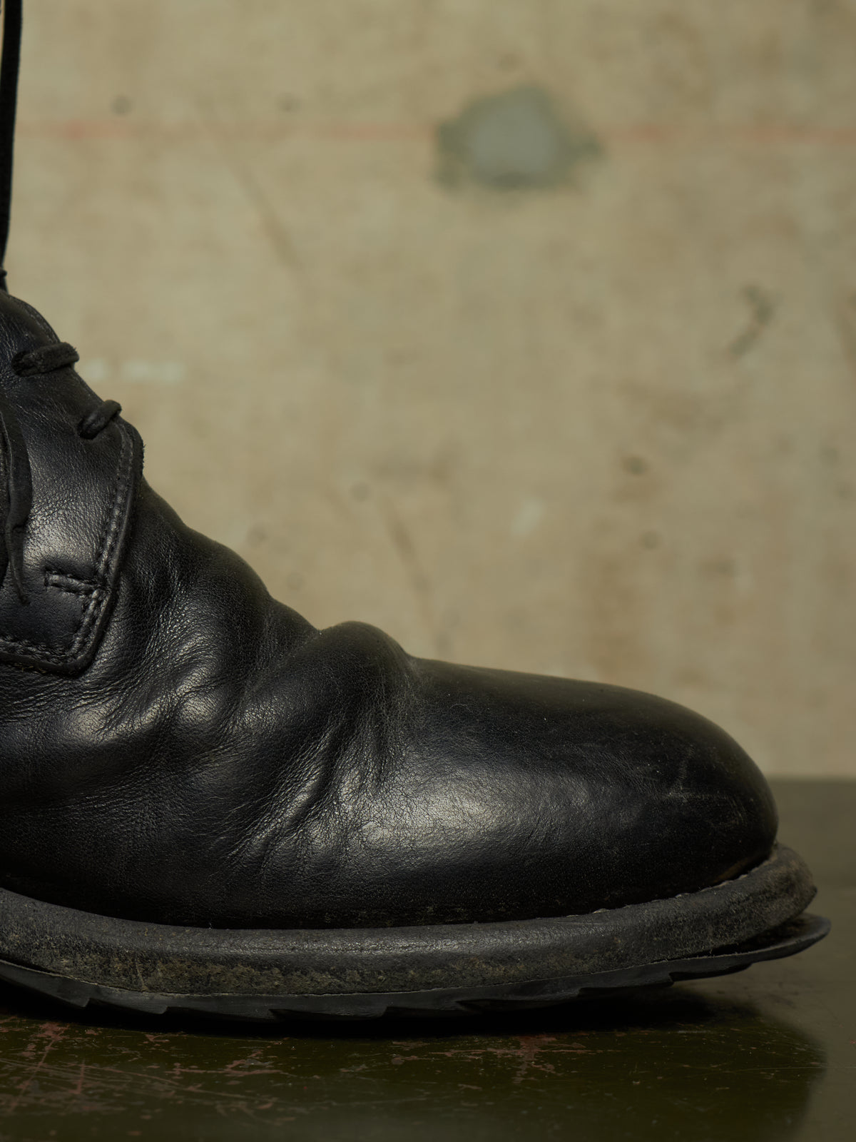 A close-up shot of a black leather lace-up boot, showing the side profile and toe, resting on a dark surface against a textured beige background.