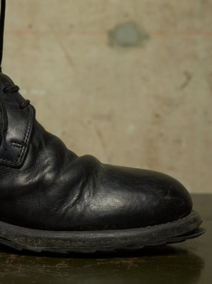 A close-up shot of a black leather lace-up boot, showing the side profile and toe, resting on a dark surface against a textured beige background.