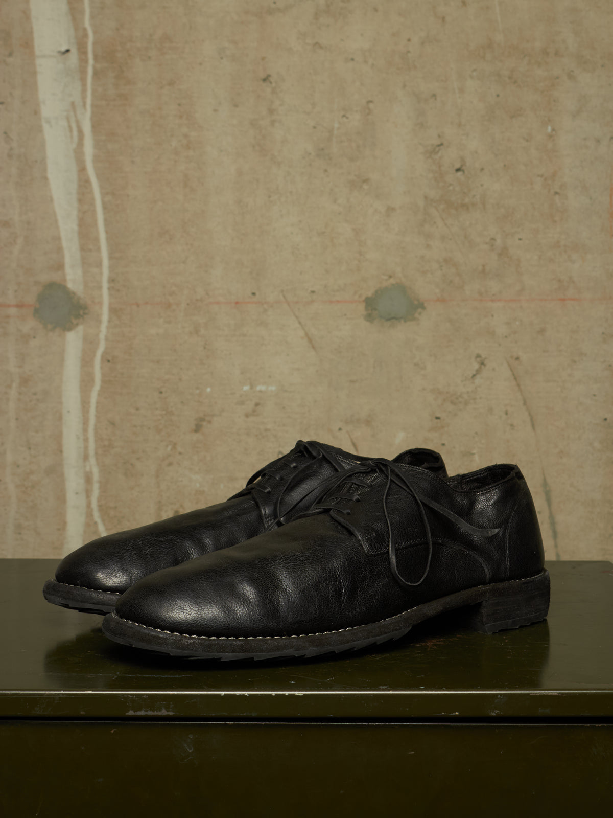 A pair of black leather lace-up dress shoes with a slightly distressed, vintage finish, displayed on a dark green surface against a textured beige wall.