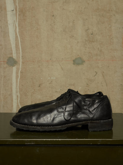 A pair of black leather lace-up dress shoes resting on a dark green surface against a textured beige wall.