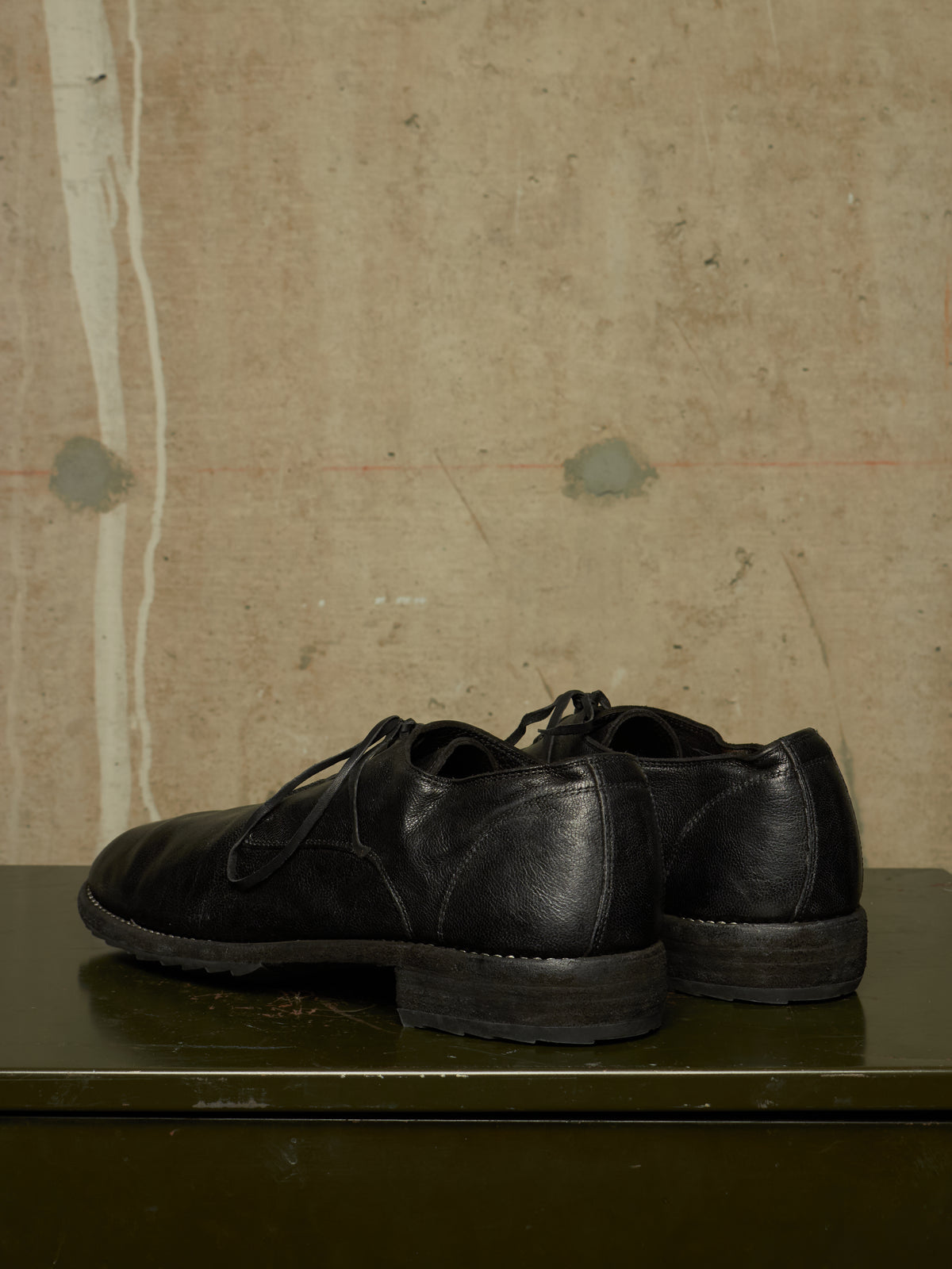 A pair of black leather lace-up dress shoes photographed from the side and back, resting on a dark green surface against a textured beige wall.