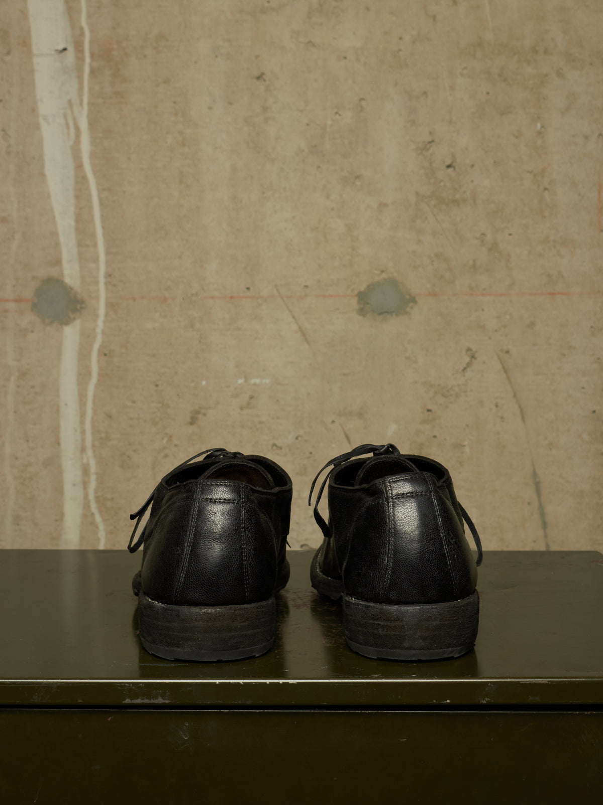 A pair of black leather lace-up shoes shown from the back, resting on a dark green surface against a textured beige wall.