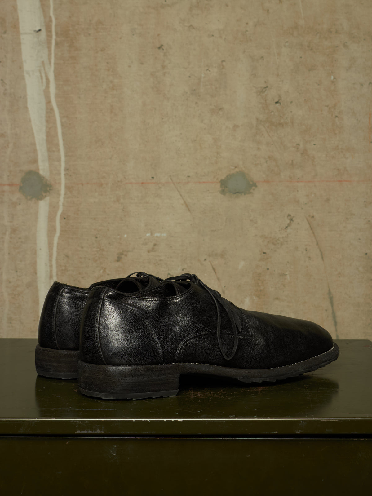 A pair of black leather lace-up dress shoes displayed on a dark green surface against a textured beige wall.