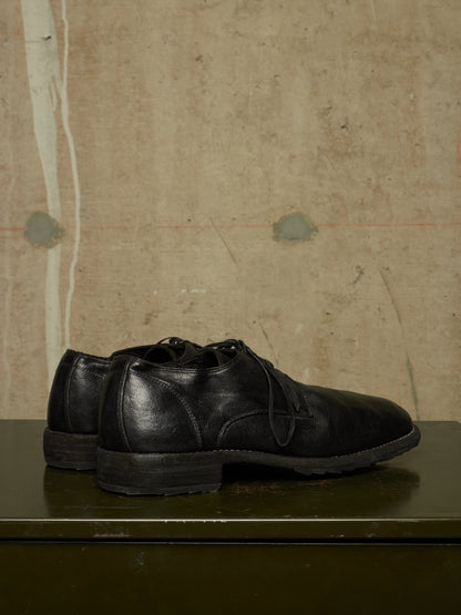 A pair of black leather lace-up dress shoes displayed on a dark green surface against a textured beige wall.