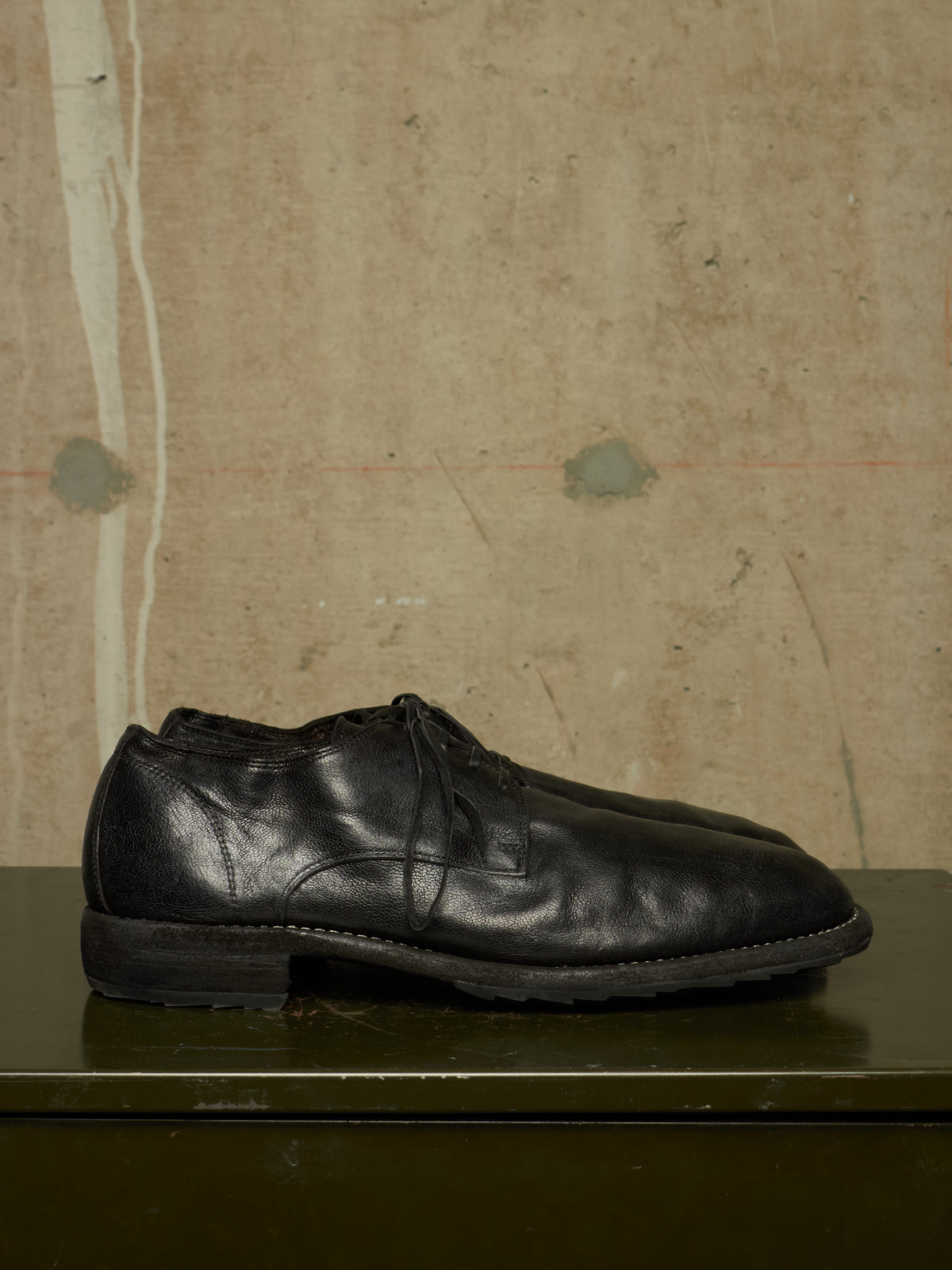 A pair of black leather lace-up dress shoes resting on a dark green surface against a textured, warm beige concrete wall.