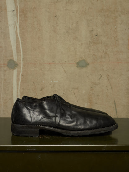 A pair of black leather lace-up dress shoes resting on a dark green surface against a textured, warm beige concrete wall.