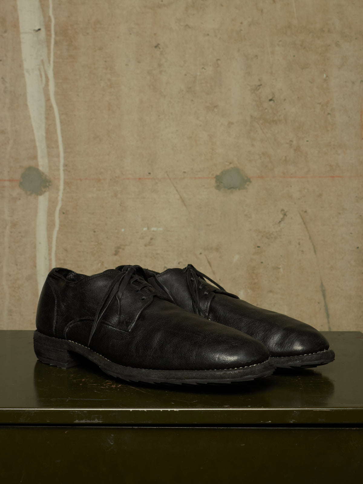 A pair of black leather lace-up dress shoes resting on a dark green surface against a textured beige wall.