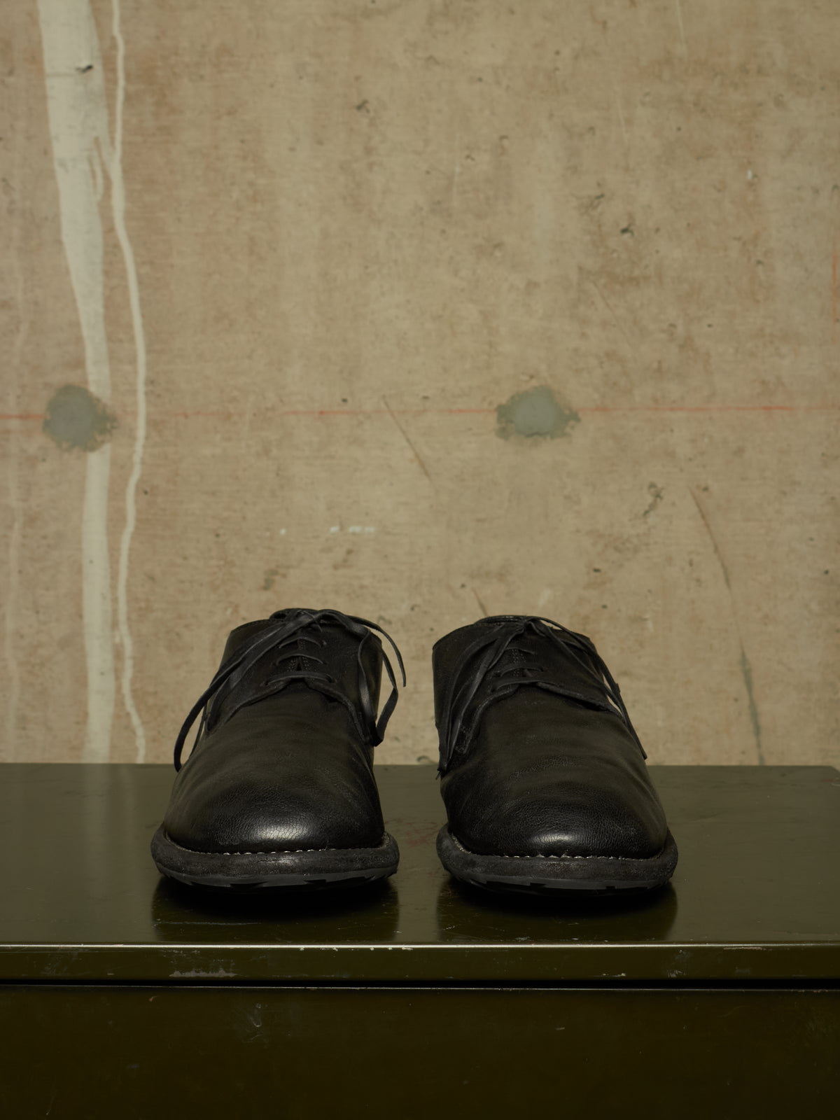 A pair of black leather lace-up shoes displayed on a dark green surface against a textured beige wall.