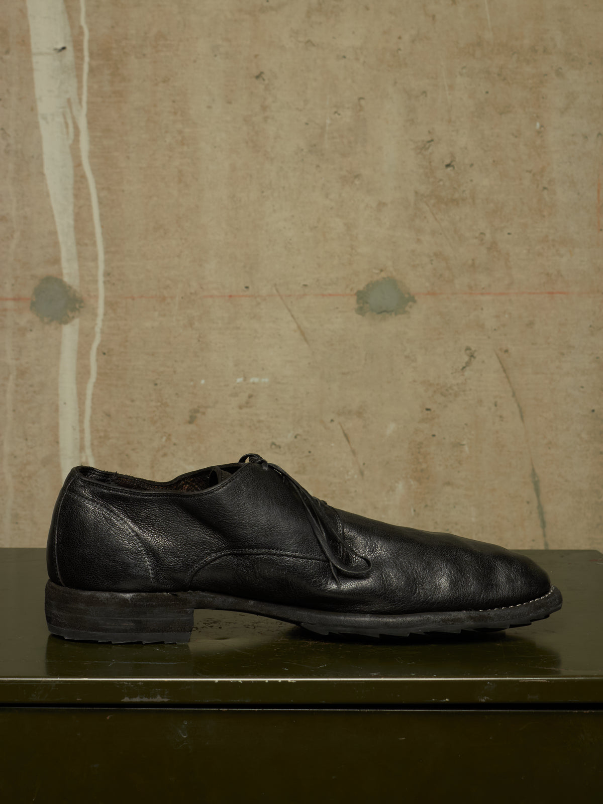 A single black leather lace-up dress shoe with a low stacked heel, resting on a dark green surface against a textured beige wall.