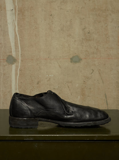 A single black leather lace-up dress shoe with a low stacked heel, resting on a dark green surface against a textured beige wall.