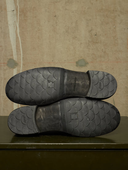The soles of a pair of black shoes, showing a textured tread pattern and the brand name 'BONSAI' printed in the center, resting on a dark green surface against a textured beige wall.