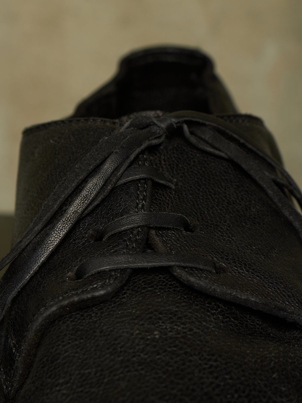 A close-up shot of a black leather lace-up shoe, focusing on the textured leather and laces against a neutral beige background.