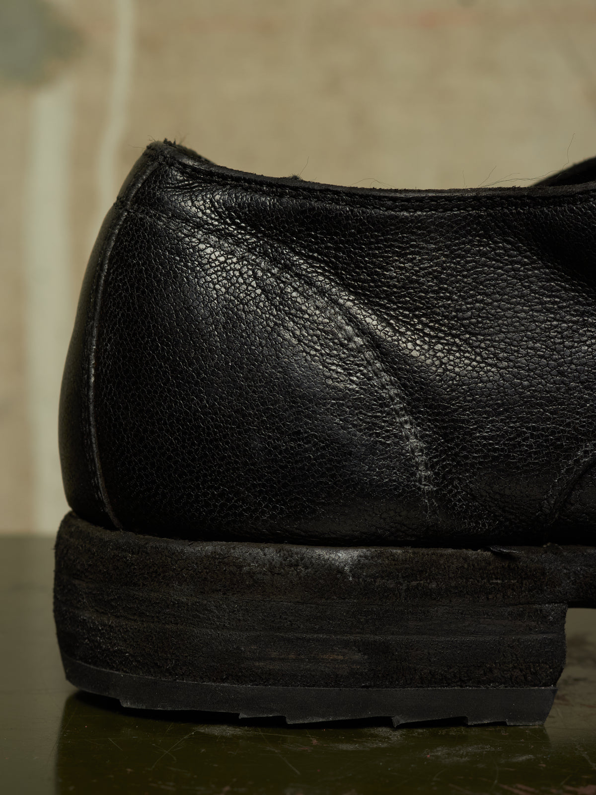 Close-up of the rear side of a black leather shoe with a chunky black sole, resting on a dark reflective surface.