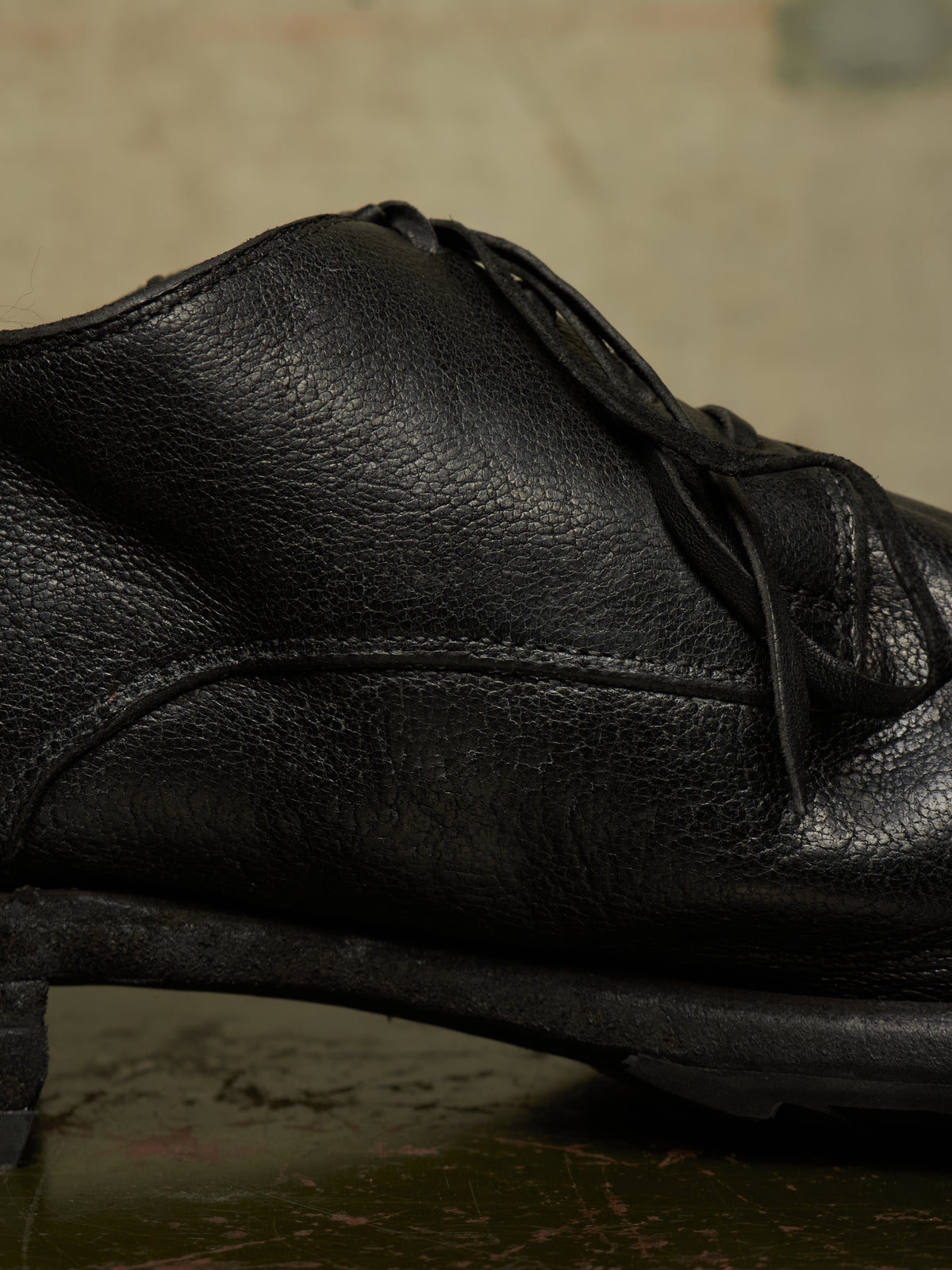 A close-up shot of a black leather lace-up shoe, showing the textured grain and stitching details against a dark, moody background.
