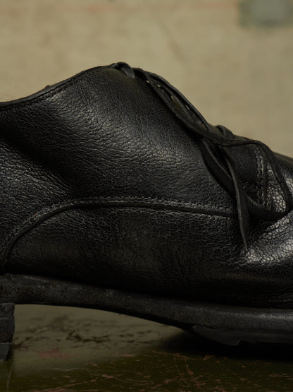 A close-up shot of a black leather lace-up shoe, showing the textured grain and stitching details against a dark, moody background.