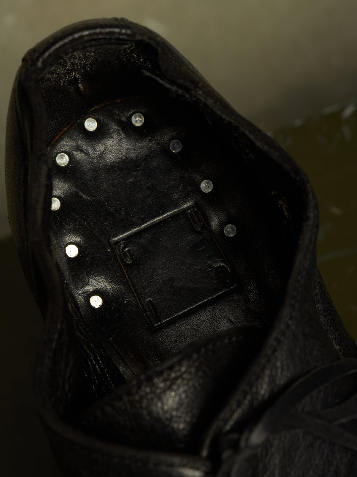 Close-up of the interior of a black leather shoe or boot, showing a black footbed with multiple small silver metal studs arranged in a circle and a rectangular black insert in the center.