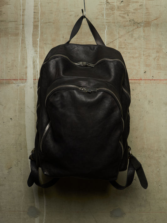 A black leather backpack with multiple zippered compartments, hanging from a metal hook against a textured concrete wall.