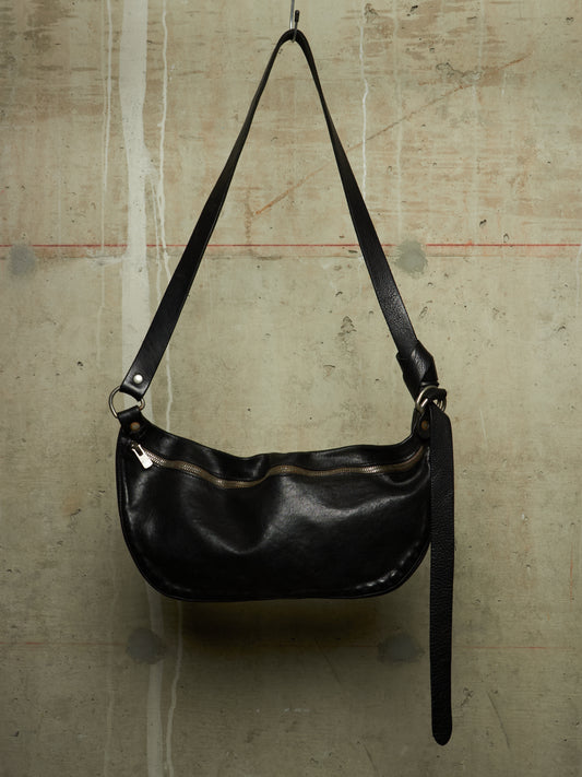 A black leather crescent-shaped shoulder bag with a long adjustable strap, hanging against a textured concrete wall.