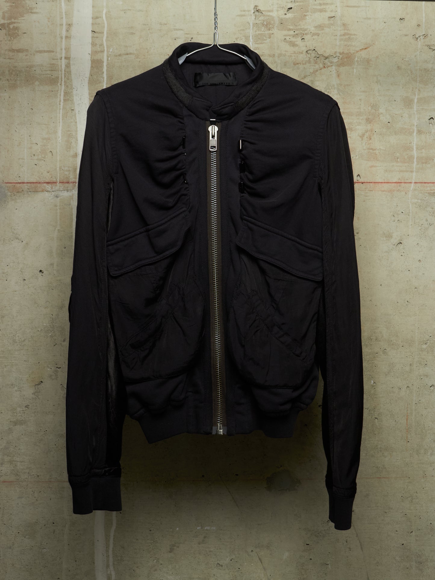 A black long-sleeve bomber jacket with a front zipper, hanging on a wire hanger against a textured concrete wall.