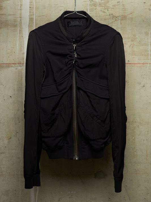 A black ruched zip-up jacket hanging on a metal hanger against a textured concrete wall.