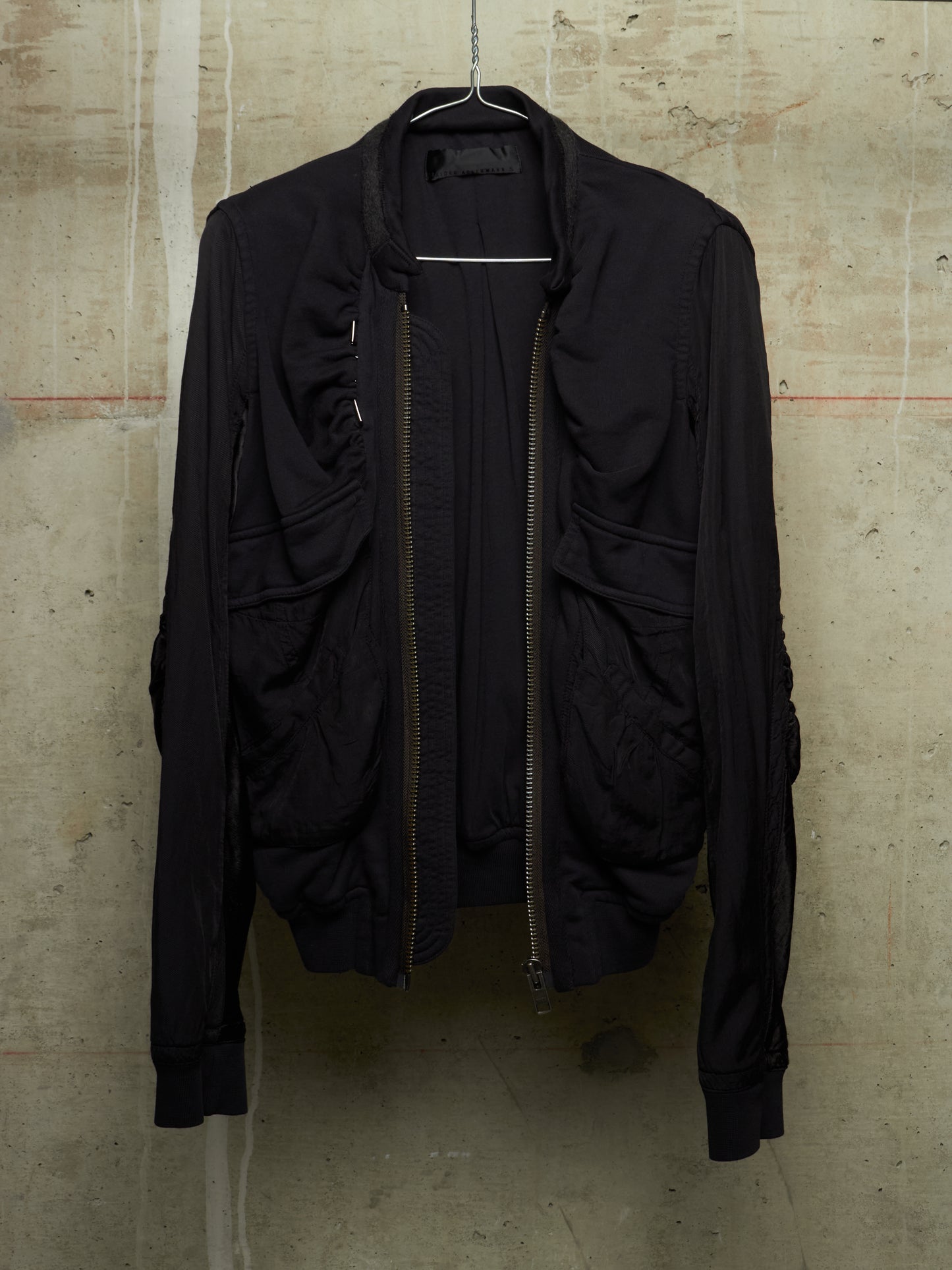 A black lightweight bomber-style jacket with a front zipper, ribbed cuffs and hem, hanging on a wire hanger against a textured concrete wall.