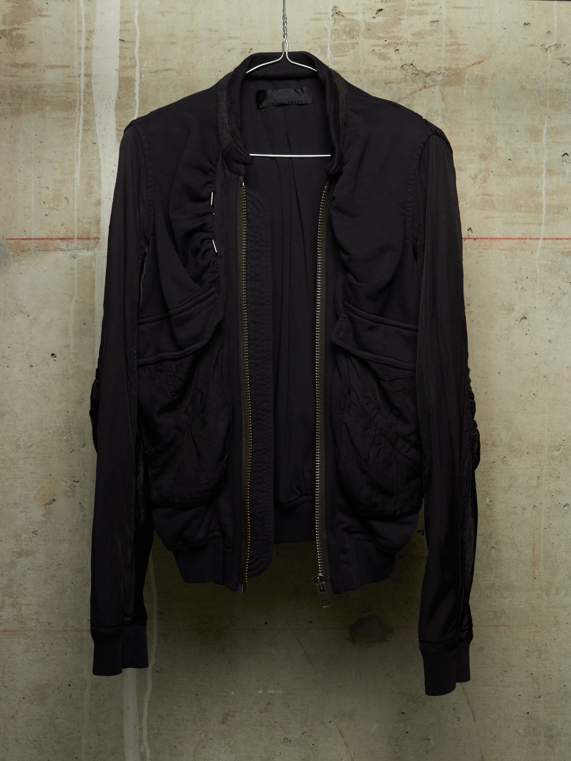 A black lightweight bomber-style jacket with a front zipper, ribbed cuffs and hem, hanging on a wire hanger against a textured concrete wall.