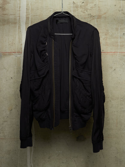 A black lightweight bomber-style jacket with a front zipper, ribbed cuffs and hem, hanging on a wire hanger against a textured concrete wall.