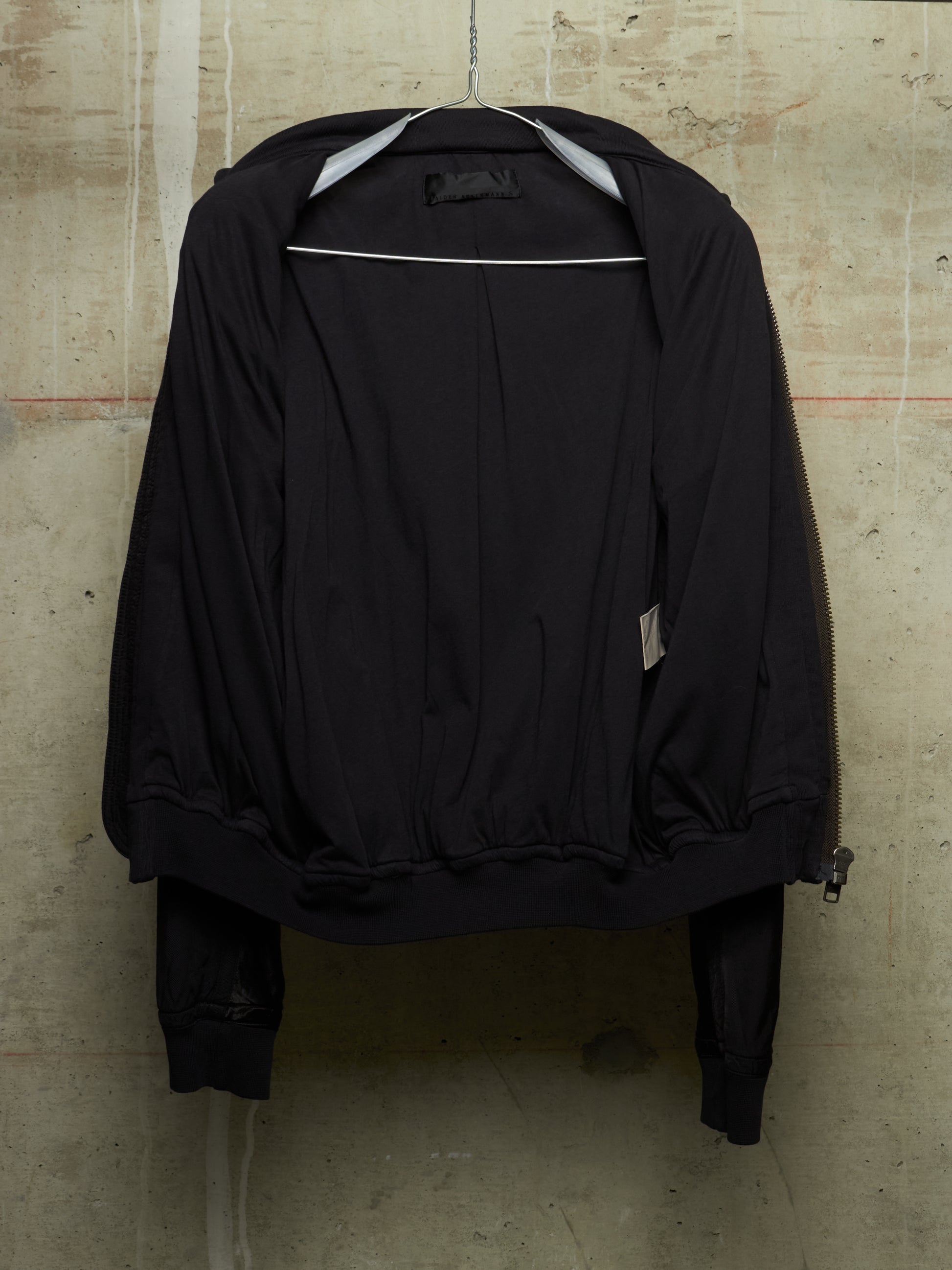 A black bomber jacket hanging on a metal hanger against a textured concrete wall. The jacket features a zippered front, elasticized cuffs and hem, and a small white brand label on the sleeve.