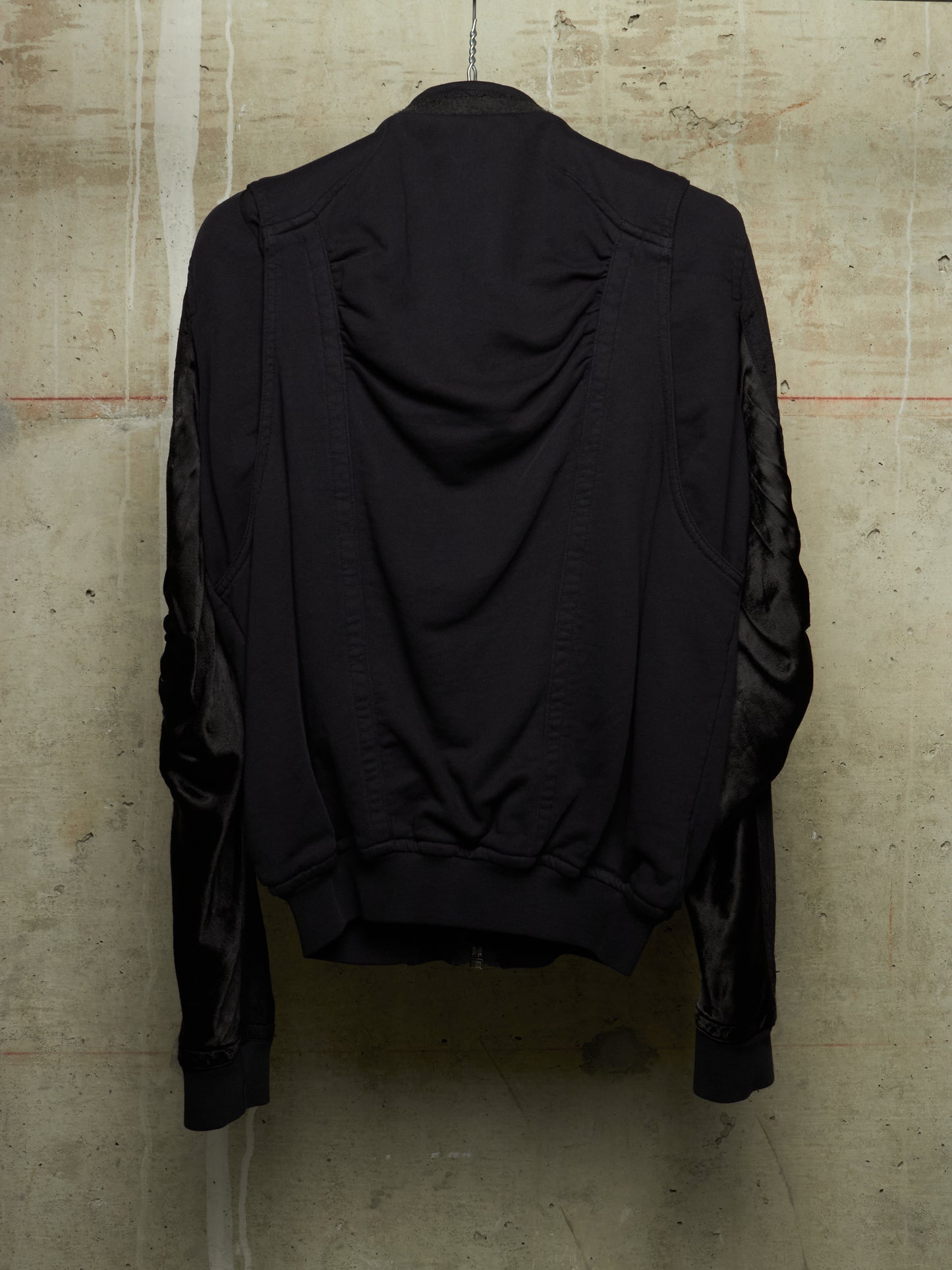 A black jacket with ruched detailing on the back and sheer black mesh sleeves, shown hanging against a textured concrete wall.