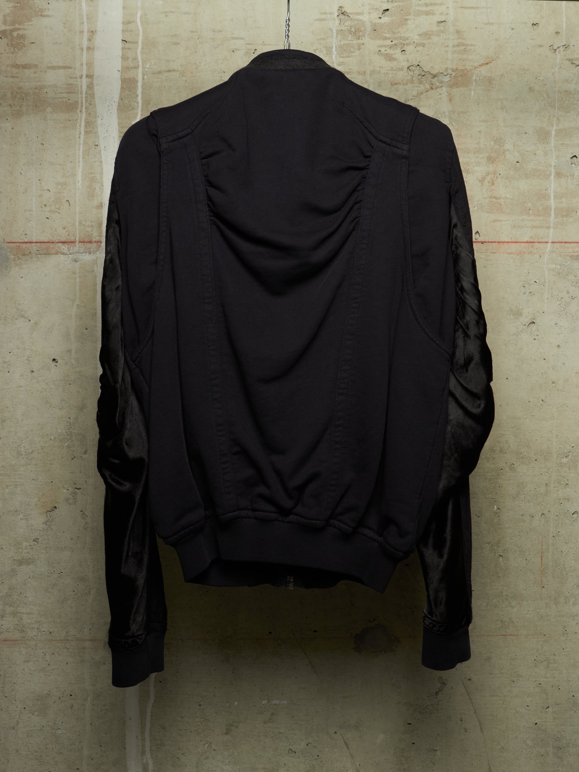 A black jacket with ruched detailing on the back and sheer black mesh sleeves, shown hanging against a textured concrete wall.