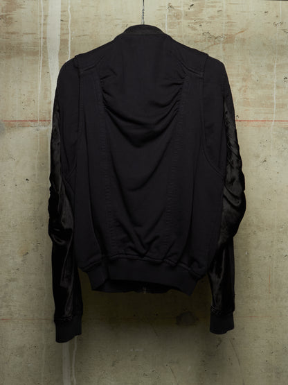 A black jacket with ruched detailing on the back and sheer black mesh sleeves, shown hanging against a textured concrete wall.