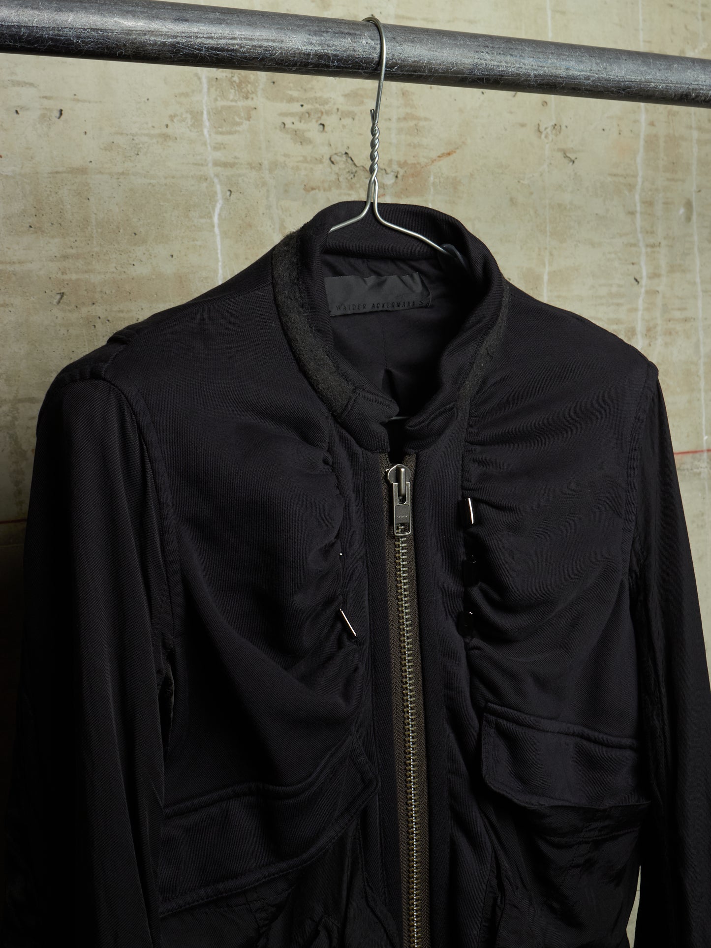 A black zip-up jacket hanging on a black hanger from a metal clothing rack, set against a textured beige concrete wall.