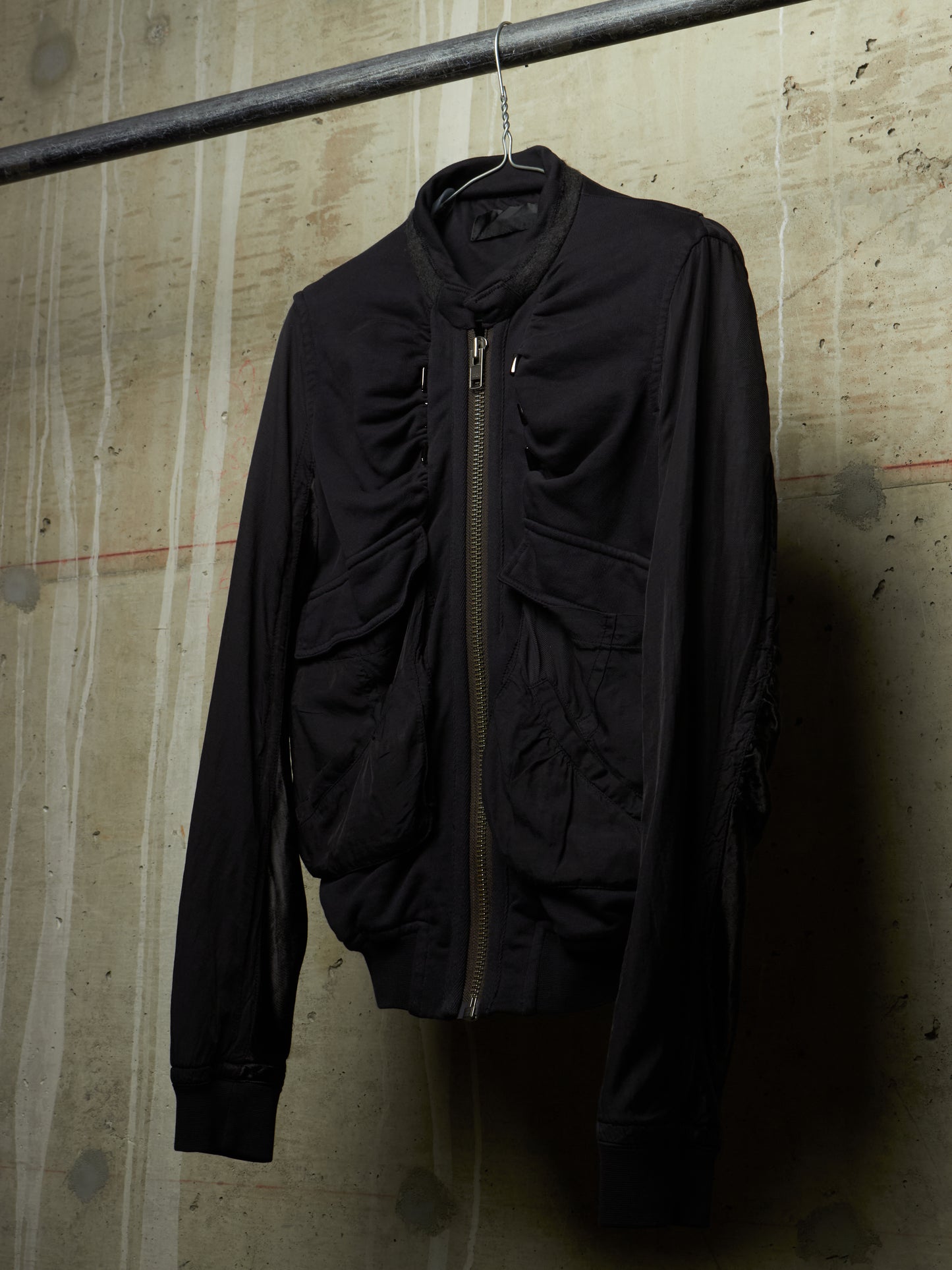 A black zip-up jacket with a high collar and multiple front pockets, hanging on a metal hanger from a horizontal rod against a textured concrete wall.