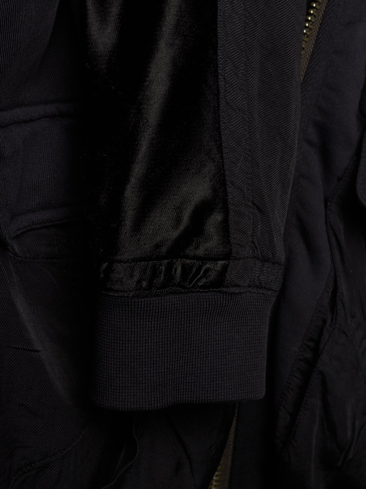 Close-up detail of a black jacket showing a ribbed fabric cuff, a satin-like black lining, and a partial gold-toned zipper.