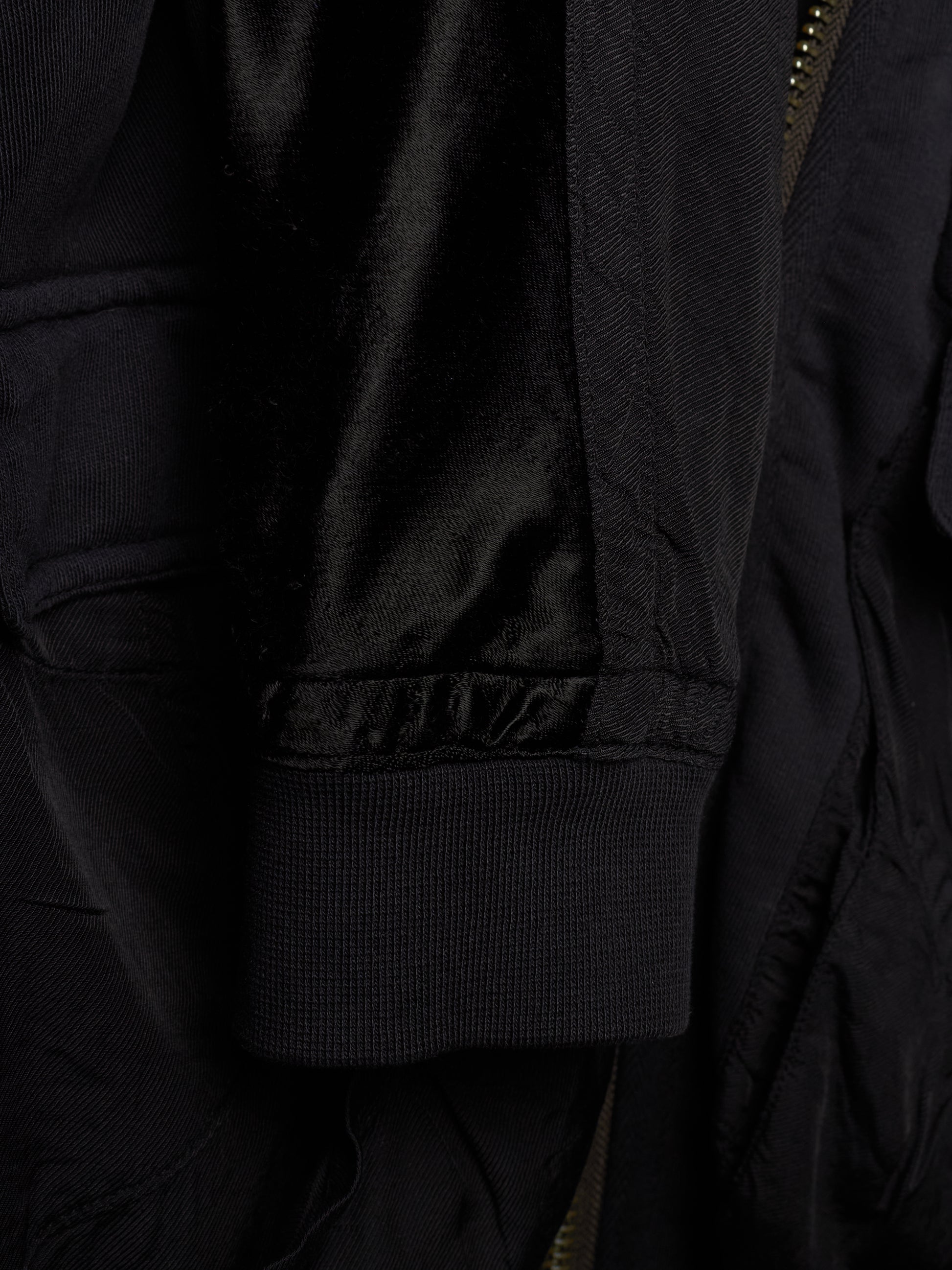 Close-up detail of a black jacket showing a ribbed fabric cuff, a satin-like black lining, and a partial gold-toned zipper.