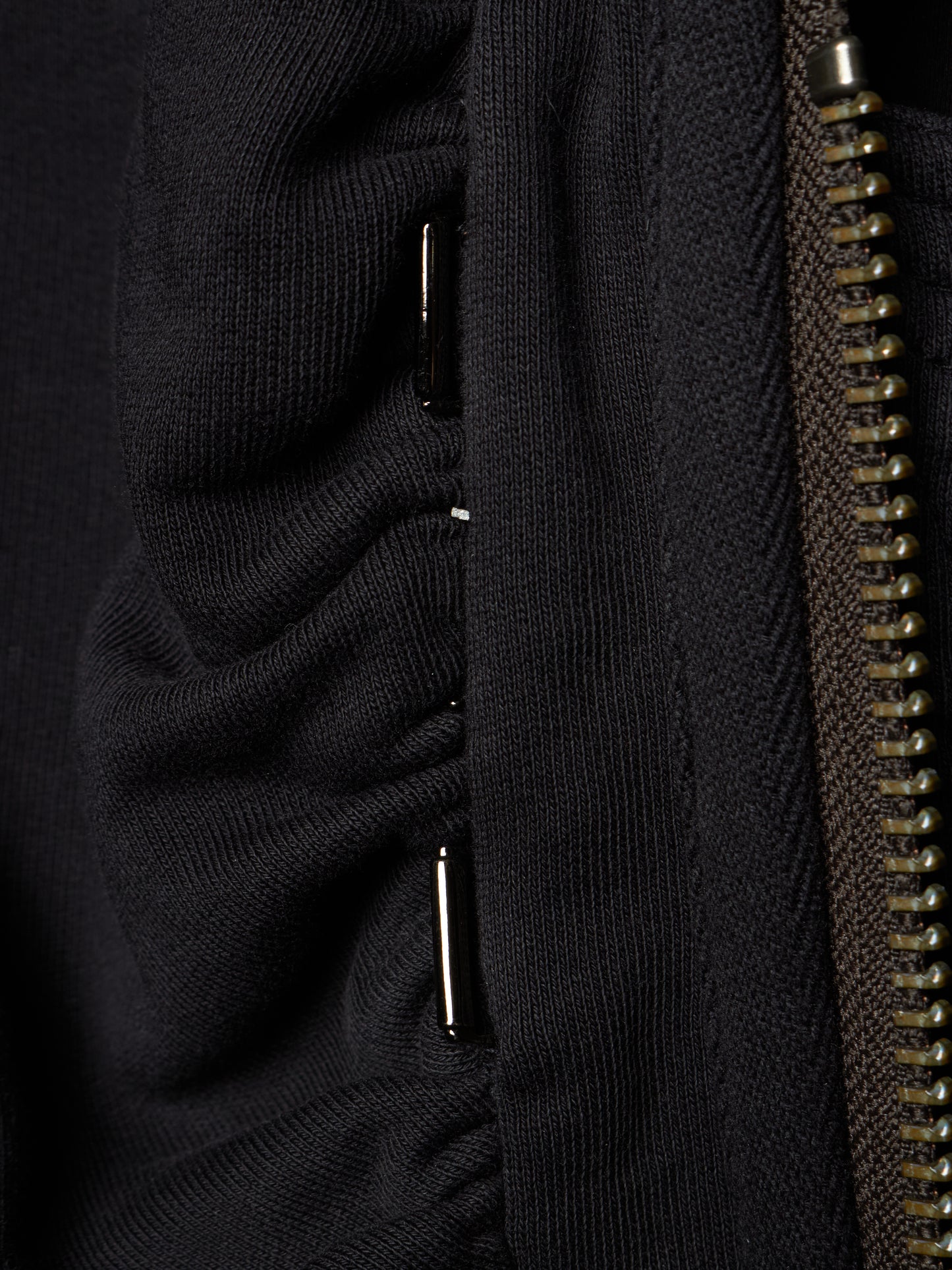 Close-up detail of a black ribbed knit garment showing a vertical seam, a metallic zipper, and small silver metal accents.