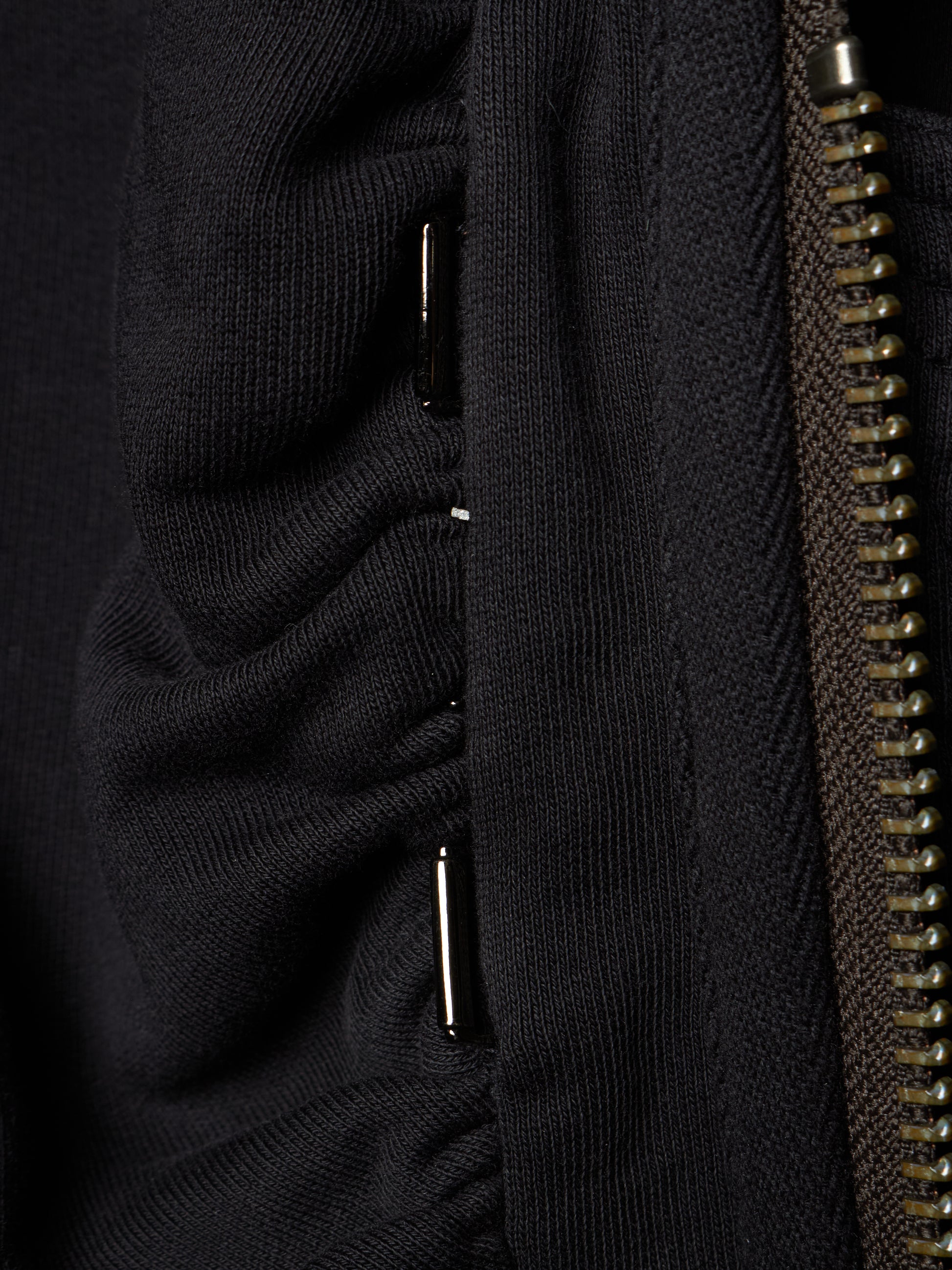 Close-up detail of a black ribbed knit garment showing a vertical seam, a metallic zipper, and small silver metal accents.