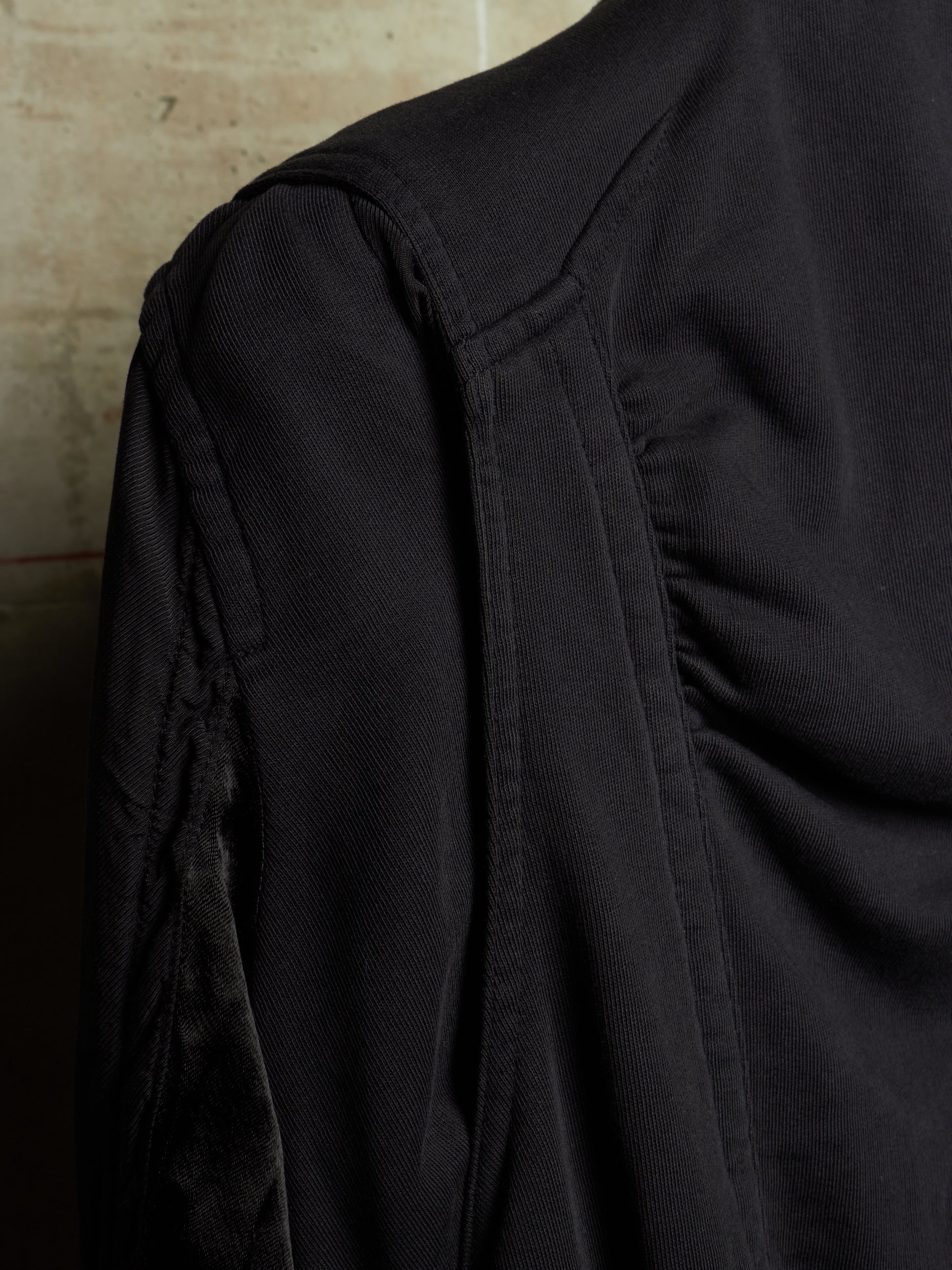 Close-up of a black textured garment, showing the shoulder seam, sleeve, and gathered fabric details against a neutral background.