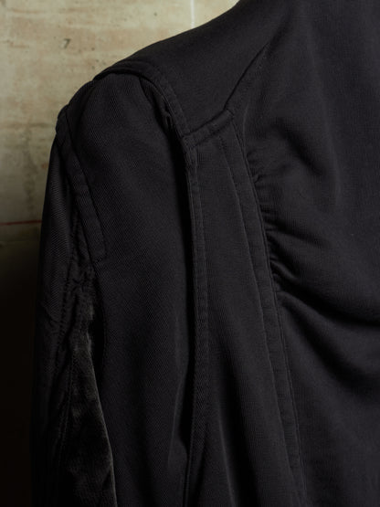 Close-up of a black textured garment, showing the shoulder seam, sleeve, and gathered fabric details against a neutral background.