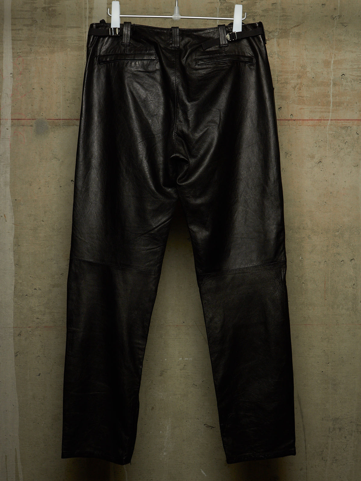 Black leather pants hanging from white clips against a textured concrete wall.