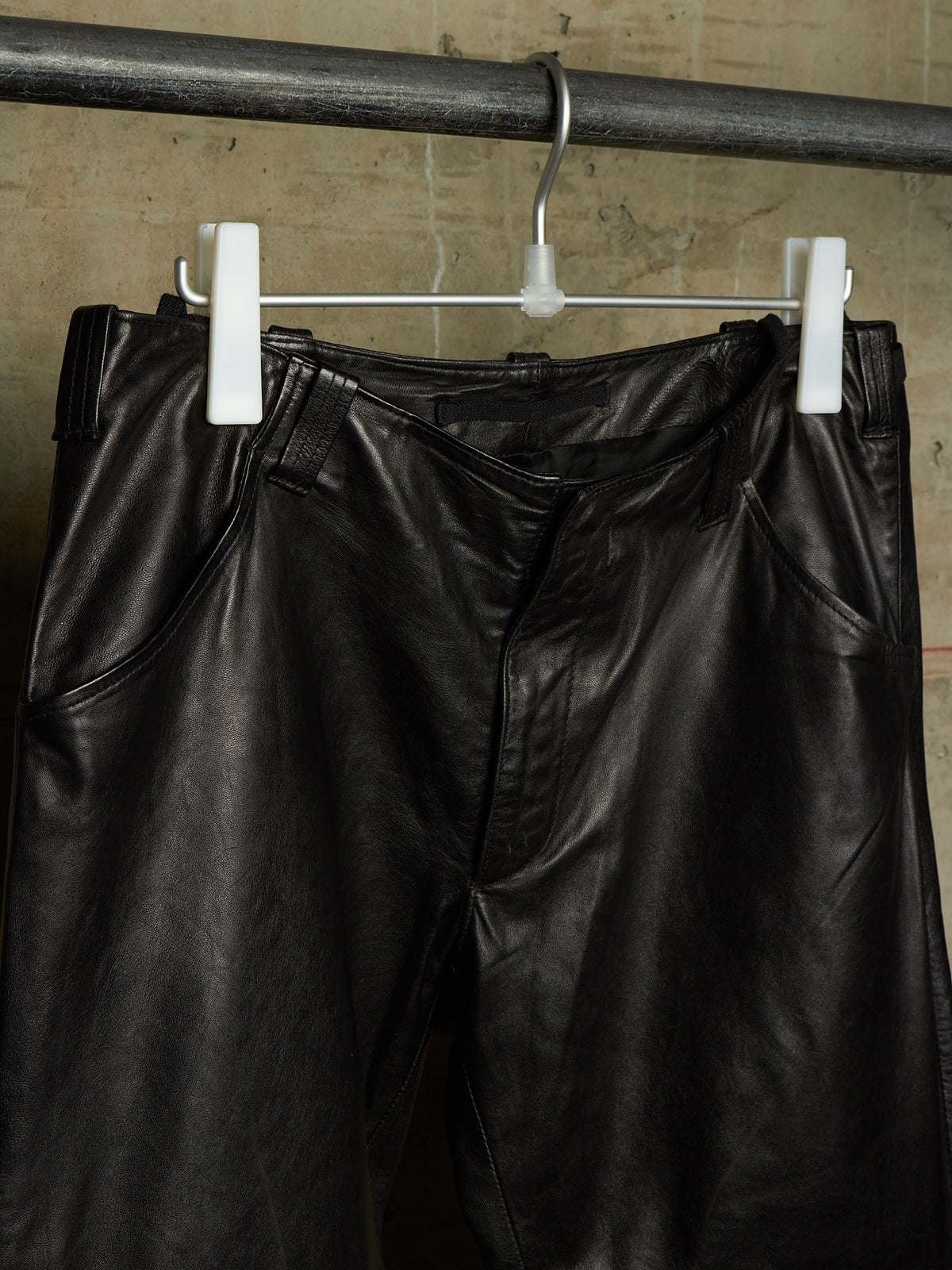 Black leather pants hanging from white plastic clips on a metal clothing rack against a textured concrete wall.