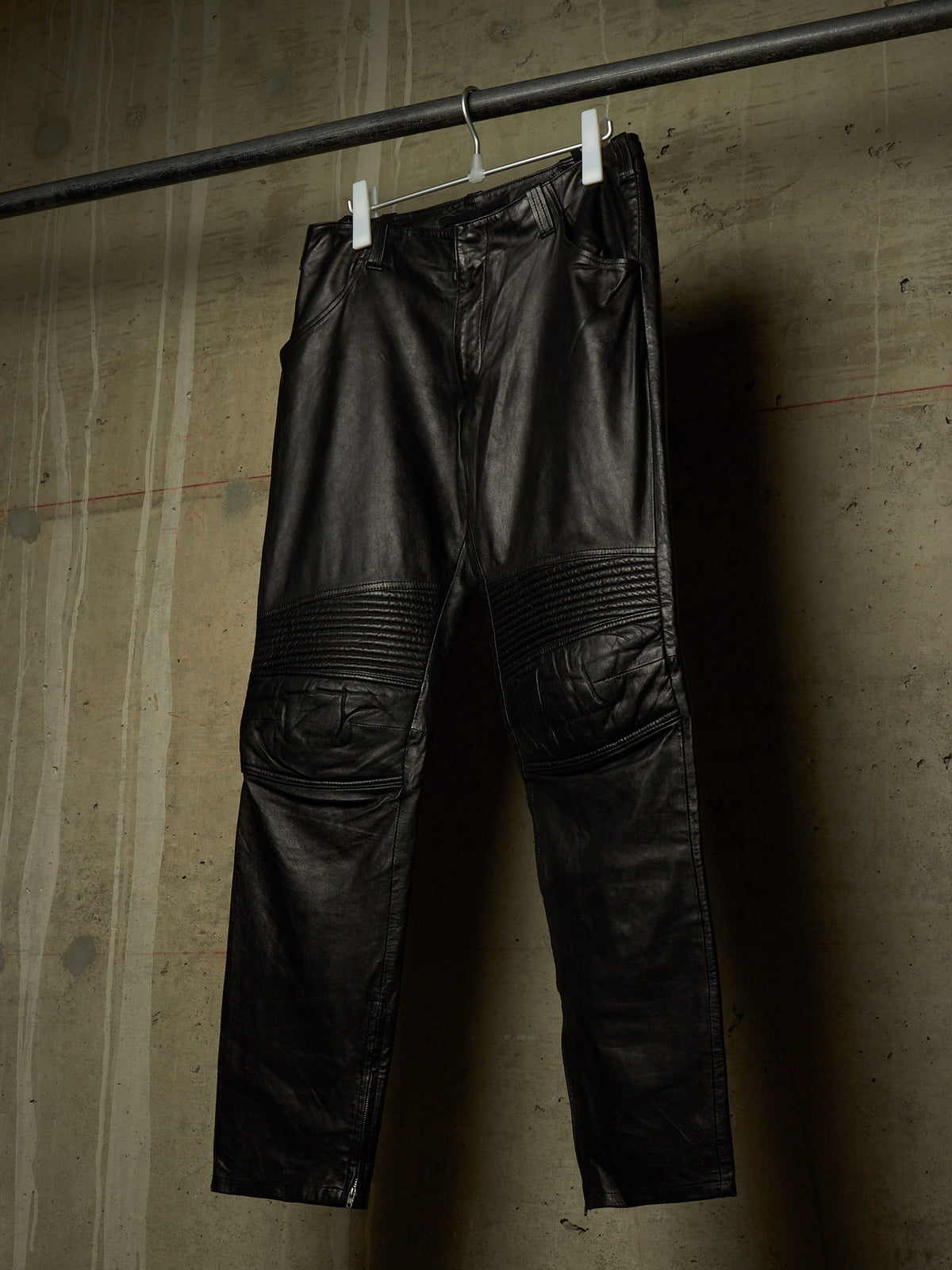 Black leather pants with ribbed knee panels, hanging from white clips on a metal rod against a textured concrete wall.