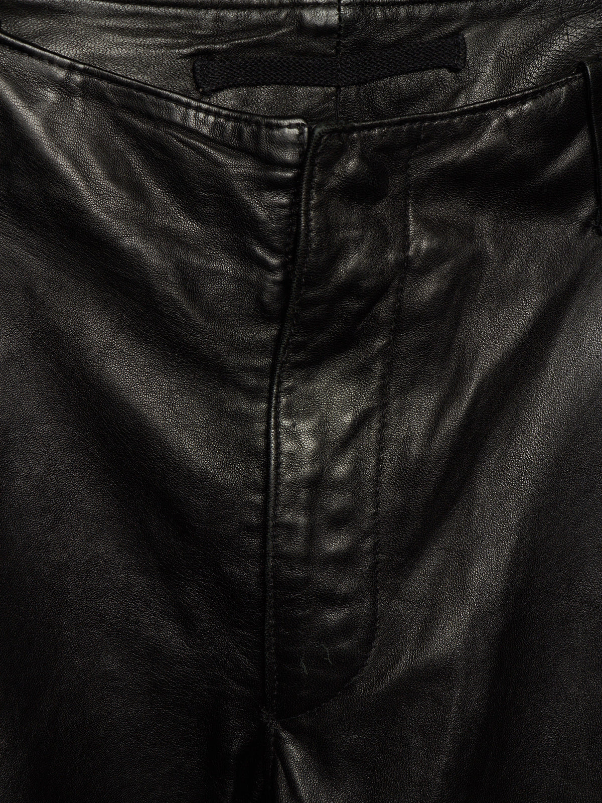 Close-up of black leather pants showing the waistband, belt loop, front seam, and texture of the material.