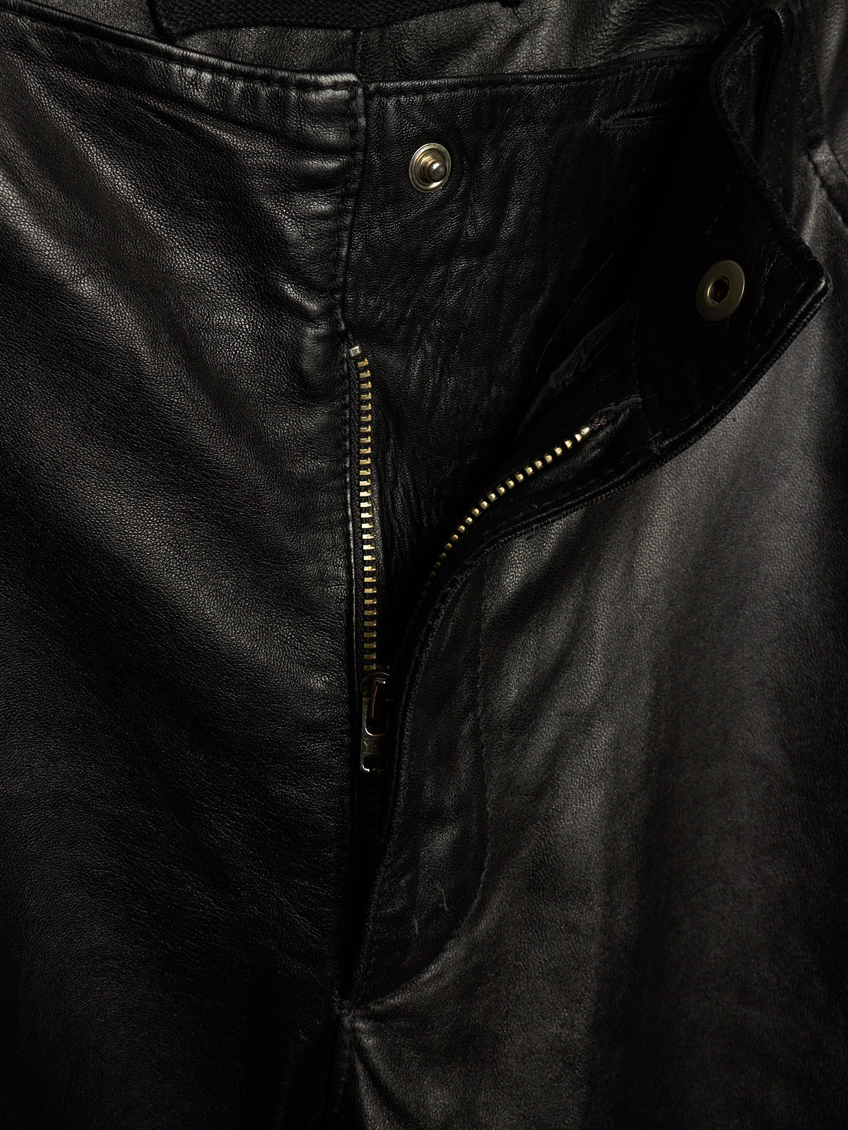 Close-up of a black leather jacket showing the front zipper, metal snap buttons, and textured leather surface.
