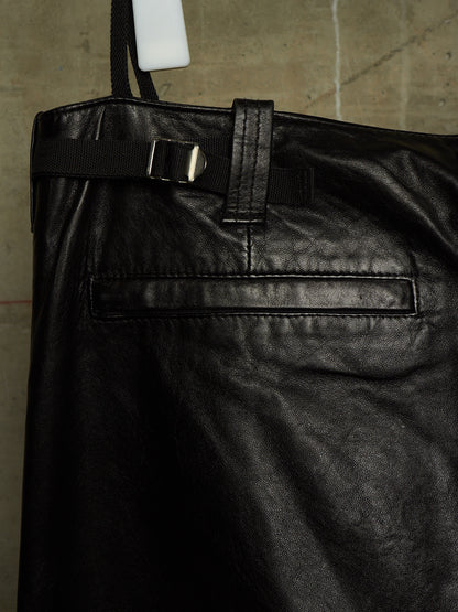 Close-up of a black leather bag or pants leg with a belt strap and silver buckle, photographed against a concrete floor background.