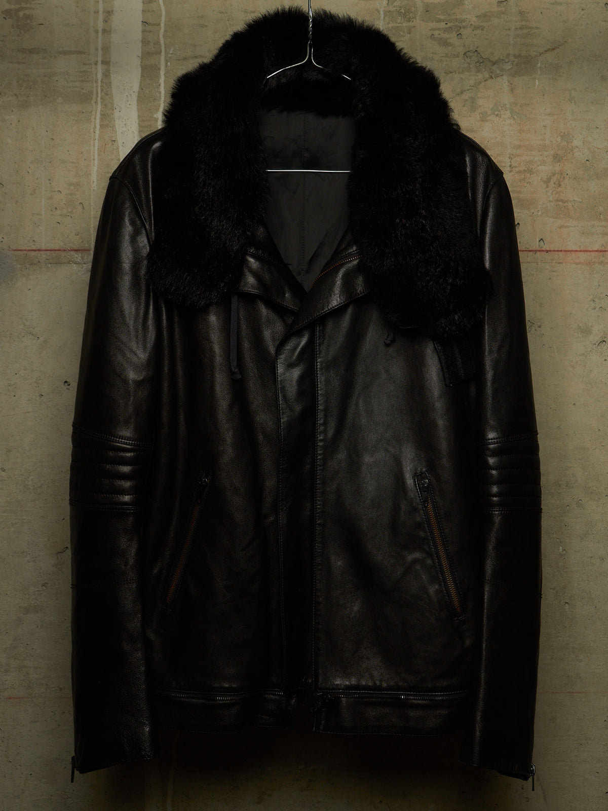 A black leather motorcycle jacket with a thick black faux-fur collar, hanging on a wire hanger against a textured concrete wall.