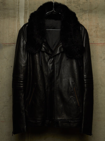 A black leather motorcycle jacket with a thick black faux-fur collar, hanging on a wire hanger against a textured concrete wall.
