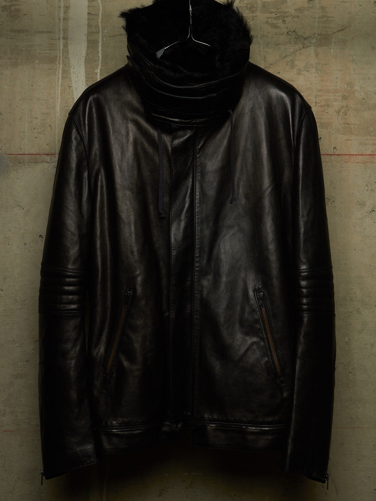 A black leather jacket with a high black faux-fur collar, hanging on a black hanger against a textured concrete wall.