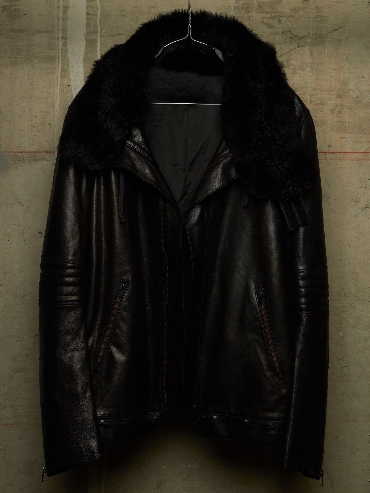 A black leather jacket with a large black fur collar, hanging on a wire hanger against a textured concrete wall.