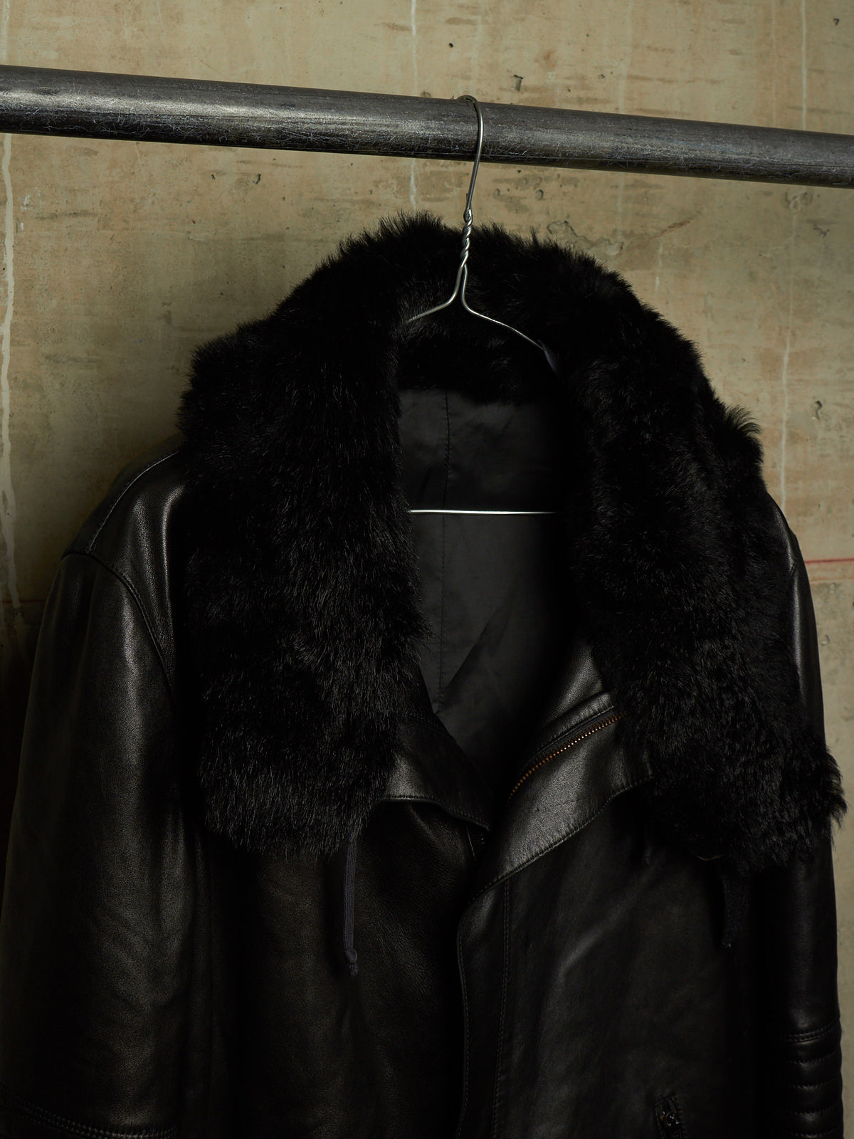 A black leather jacket with a large black fur collar, hanging on a metal hanger from a dark horizontal rod against a textured beige wall.
